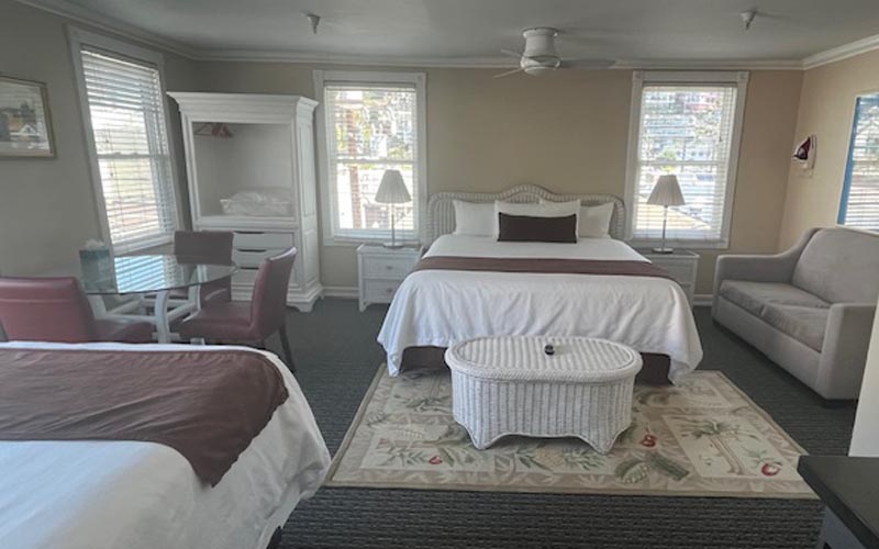 Hotel room with two white beds, wicker coffee table, nightstands, and a beige sofa in a bright space with windows on both sides