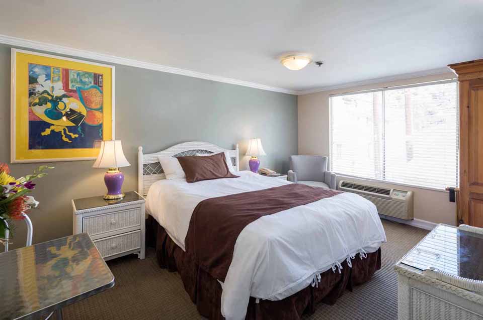 Hotel room with a white bed, brown bed runner and pillows, flanked by matching nightstands and purple lamps
